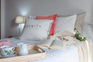 cama con cojines blancos y rosa, bandeja de madera con desayuno y lámpara encendida